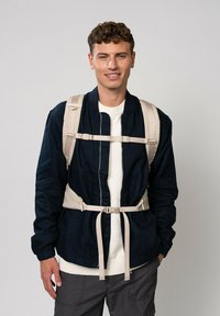 Veste légère navy avec des sangles et des accessoires beiges, portée sur un pull blanc. Un pantalon gris avec des poches termine la tenue.