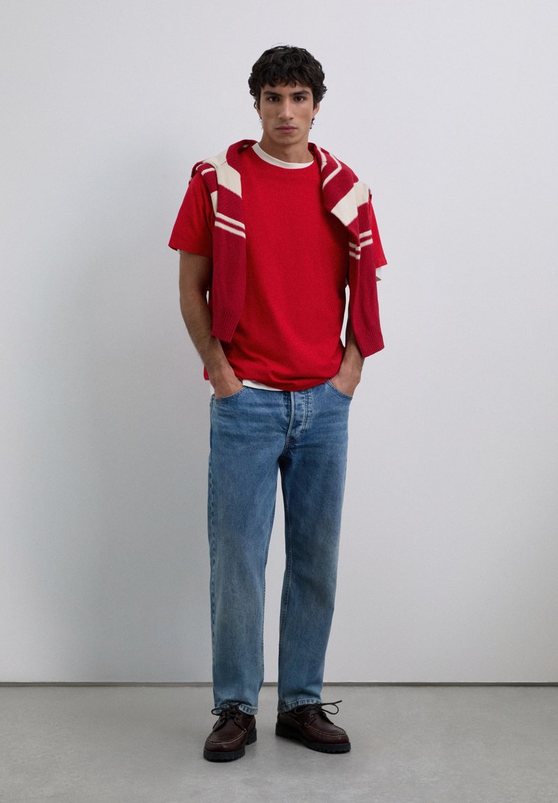 Jonge man staat met handen in zijn zakken, draagt een rood T-shirt, een gestreepte trui over zijn schouders, blauwe jeans en donkerbruine schoenen.