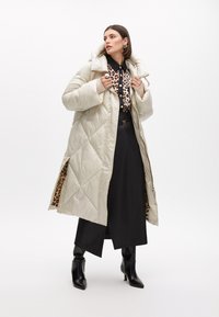 Vadderad krämfärgad jacka med diamantquiltning, med en pälskantad huva och leopardmönstrad foder. Bärs över en svart outfit och ankelstövlar.