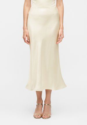 Femme portant une jupe midi en satin crème et des talons ouverts dorés à bride à la cheville, debout devant un fond blanc.