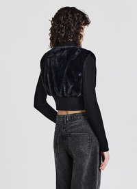 Schwarzer, taillierter Pullover mit einem plüschigen Pelzrücken und gerippten Ärmeln, kombiniert mit grauen, hochgeschnittenen Jeans. Glattes Texturdetail sichtbar.