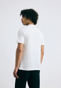 T-shirt blanc à manches courtes avec un col rond, texture douce, coupe ajustée, porté avec un pantalon texturé foncé, modèle tourné dos à l'appareil.