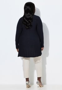 Kvinde med langt sort hår iført en navyblå ribstrikket lang sweater, cremefarvede cropped bukser og høje sko med slangeskindsprint, stående op af en hvid væg.