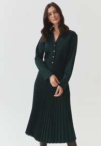 Abito lungo a maniche lunghe di colore verde scuro con scollatura a V, dotato di bottoni dorati e design a gonna plissettata. Texture morbida su tutto l'abito.