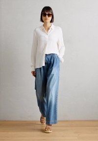 Chemise blanche à boutons, pantalon en denim bleu clair ample, sandales marron et lunettes de soleil foncées. Design minimaliste et coupe décontractée.