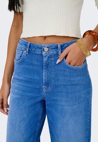 Šviesiai mėlyni aukštos liemens džinsai su lygia tekstūra, penkių kišenių dizainu ir sidabrine sagos sagtimi, derinami su baltu, ribotu viršumi.