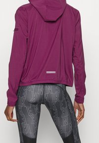 Veste légère à capuche de couleur violet foncé, dotée de poignets élastiques et d'un ourlet à cordon de serrage. Associée à des leggings noirs à motifs affichant un logo.
