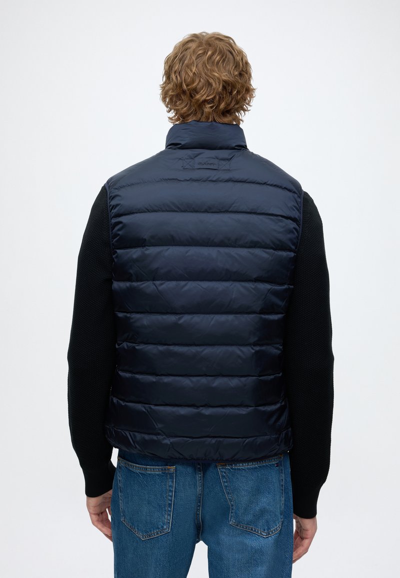Mand iført en mørkeblå quiltet vest over en sort sweater og blå jeans, set bagfra mod en ensfarvet baggrund.