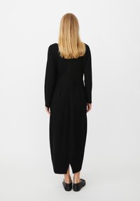 Czarna sukienka z długimi rękawami i luźnym krojem, z rozcięciem z tyłu i plisowanymi detalami, wykonana z miękkiej, gładkiej tkaniny.