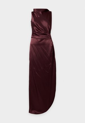 Femme debout portant une robe en satin bordeaux foncé sans manches avec une fente haute et des talons aiguilles noirs sur fond blanc.