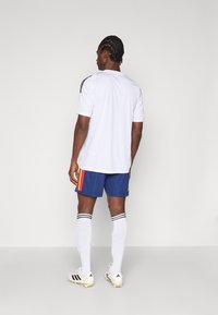 Ανδρική ποδοσφαιρική στολή που περιλαμβάνει ένα λευκό μπλουζάκι κοντομάνικο, ναυτικά σορτς με κόκκινες και κίτρινες ρίγες, λευκές κάλτσες μέχρι το γόνατο και παπούτσια με τακούνια.