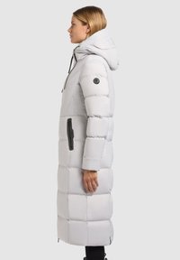 Lang hvid pufferjakke med hætte, der har et quiltet design, lynlåslommer, justerbare snore og sorte detaljer.