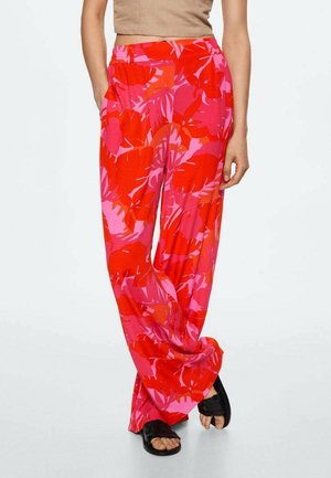 Kvinde iført løstsiddende røde og pink bukser med blomsterprint og sorte sandaler, stående med den ene hånd i lommen.