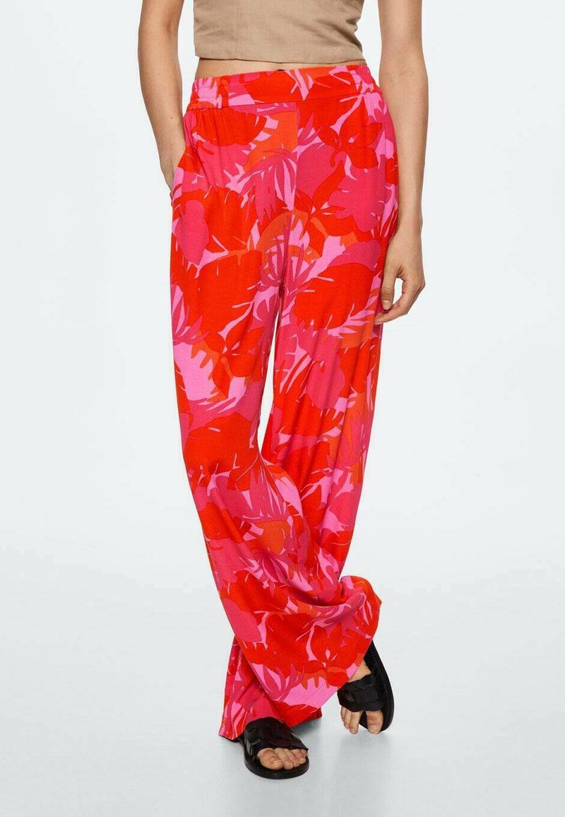Femme portant un pantalon ample à imprimé floral rouge et rose et des sandales noires, debout avec une main dans la poche.