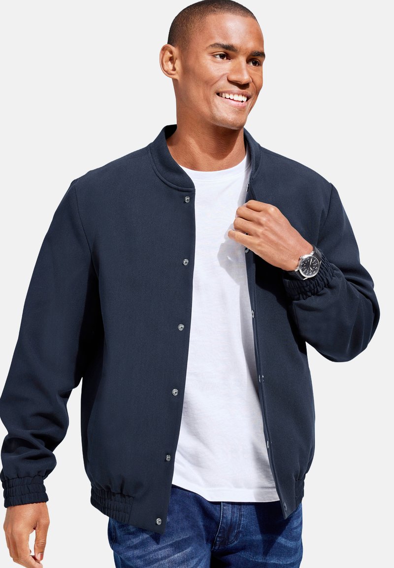 Navy bomber jacket with a round neckline, snap buttons, ribbed cuffs, and a smooth texture, worn over a white t-shirt and blue jeans.