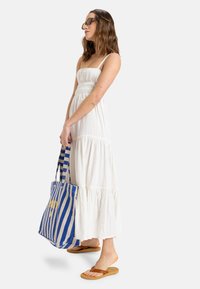 Witte, mouwloze maxi-jurk met geplooide bodice en gelaagde rok. Vrouw draagt een blauw-witte gestreepte totebag met gouden accenten. Platte sandalen.