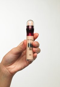 Correttore Maybelline in un tubo trasparente con un liquido rosa-beige, dotato di un applicatore a cupola trasparente e di un cappuccio rosso scuro.