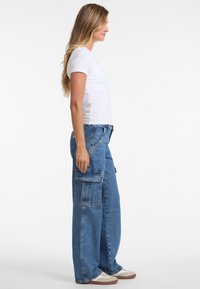 Weißes T-Shirt mit kurzen Ärmeln, kombiniert mit lockeren blauen Denim- Cargo-Hosen mit großen Seitentaschen und einem entspannten Schnitt, getragen mit hellbraunen Sneakers.