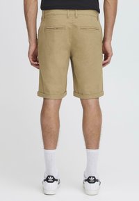 Pantalones cortos de algodón beige con un corte recto, dobladillos con vuelta y dos bolsillos traseros, llevados con calcetines blancos y zapatillas de deporte en blanco y negro.
