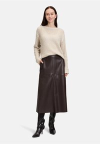 Maglione lavorato a maglia beige con vestibilità ampia, abbinato a una gonna midi in pelle marrone scuro e stivali neri sopra il ginocchio, in piedi davanti a uno sfondo neutro.