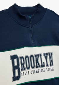 Felpa blu navy con cerniera, collo a coste e una striscia orizzontale bianca con la scritta "BROOKLYN STATE CHAMPIONS LEAGUE" in grassetto lettere nere.