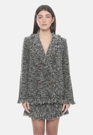 Donna che indossa una giacca doppiopetto nera e bianca testurizzata con frange e gonna coordinata con frange, in piedi su uno sfondo chiaro semplice.