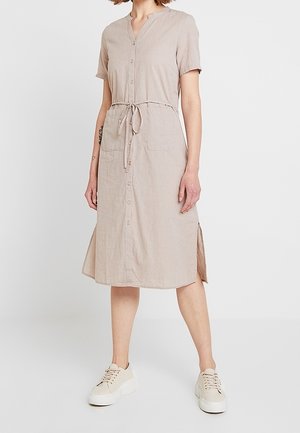 Nainen, joka pukeutunut beigeen, napilliseen midimekkoon, jossa on lyhyet hihat, vyötärönauha, etutaskut, sivuaukot ja valkoiset korotetut tennarit.