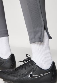 Cizmă de fotbal neagră cu o suprafață texturată și logo alb Nike, asortată cu șosete atletice albe și pantaloni de trening gri, ajustați, cu fermoare.