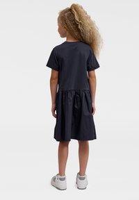 Marineblaues Kleid mit tailliertem Oberteil und gerafftem Rock, aus Baumwolle. Kurze Ärmel, knielang und ein schlichtes Design. Getragen mit weißen Sneakern.