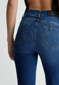 Tmavě modré džíny z denimu s vysokým pasem, dvěma zadními kapsami a hladkou texturou. Obsahují detail loga na černém pásku.