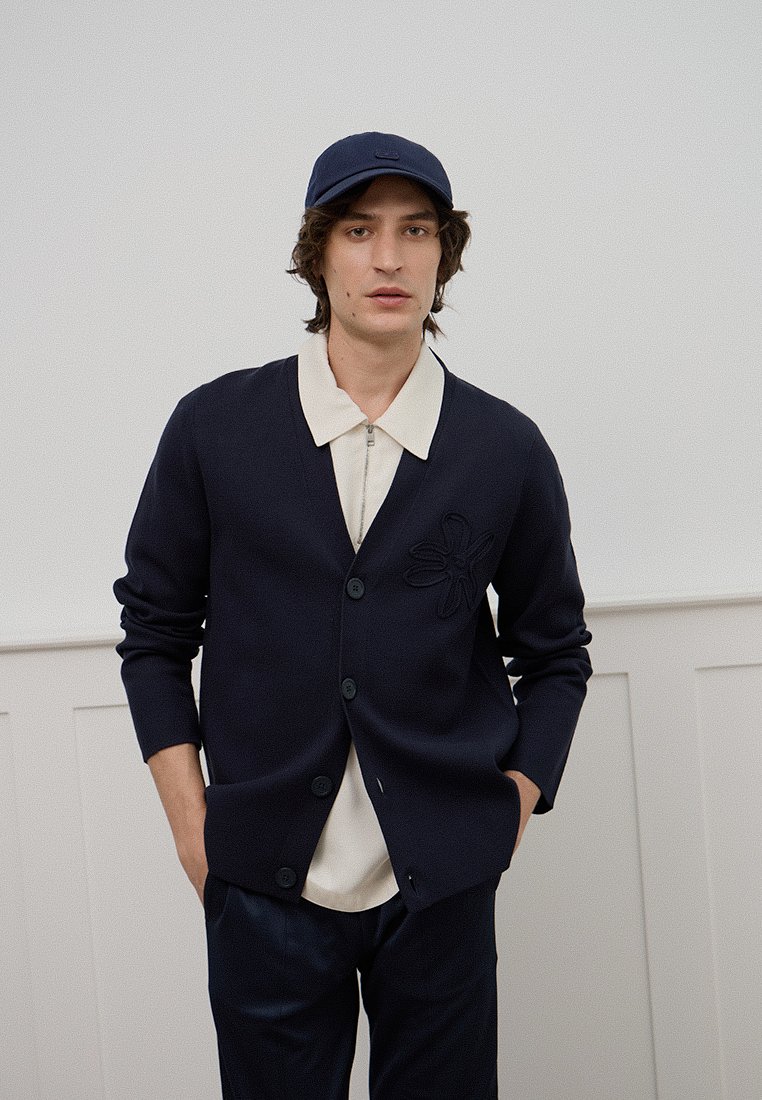 Marineblauer Cardigan aus Strickmaterial mit floralem Muster-Akzent auf der linken Seite, kombiniert mit einem hellen Hemd mit Kragen und dunklen Hosen.