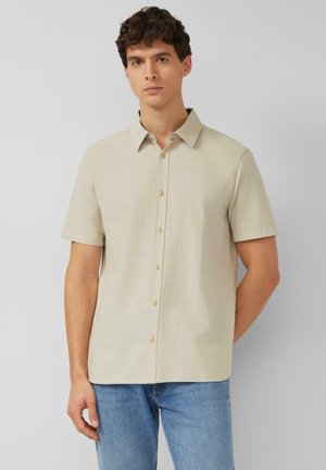 Homme portant une chemise beige à manches courtes boutonnée et un jean bleu, debout avec une main derrière le dos sur un fond uni.
