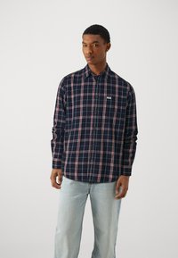 Camicia a plaid navy con motivo a quadri rossi e bianchi, maniche lunghe e una piccola tasca sul petto; abbinata a jeans azzurri chiari.