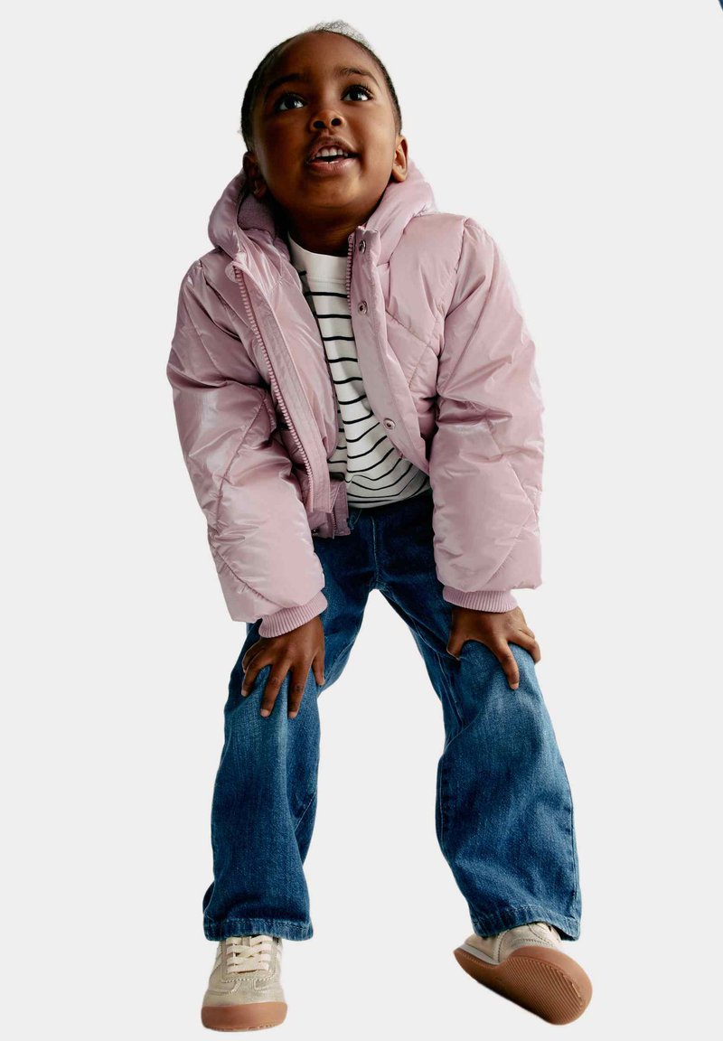 Chaqueta acolchada rosa claro con capucha, cierre de cremallera y puños acanalados, combinada con una camiseta a rayas y jeans azules. Zapatillas crema con suelas marrones.