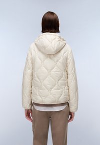 Giacca a vento imbottita in crema con cappuccio, caratterizzata da un motivo a cuciture ondulate e orlo elastico. Indossata sopra un top marrone a strati e pantaloni beige.
