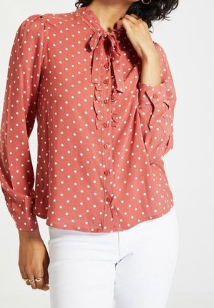 Blusa a maniche lunghe di colore ruggine con pois bianchi, con una parte anteriore arricciata e un fiocco al colletto. Polsini con bottoni e vestibilità comoda.