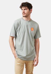 Camiseta de algodón gris claro con cuello redondo, mangas cortas y un logotipo circular rojo y amarillo en el pecho. Diseño sencillo, ajuste relajado.