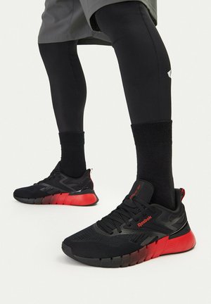 Osoba ubrana w czarne i czerwone buty sportowe Reebok, czarne skarpetki i czarne legginsy, ustawiona do chodzenia lub biegania.