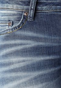 Blå denimjeans med en blekt yta, med gula sömmar och knäppning. Tyget har en slät textur och synliga vävmönster.