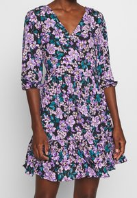 Blumenkleid mit V-Ausschnitt, dreiviertel Ärmeln und rüschengesäumtem Saum. Hat einen schwarzen Hintergrund mit lila und türkisen Blumen. Aus Baumwolle.