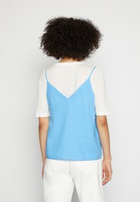 Camisola azul-clara com alças finas, costas em V, sobre uma blusa de mangas curtas brancas. Tecido suave, design minimalista.