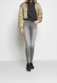 Chaqueta acolchada oliva claro, cima negra con cremallera, jeans ajustados grises y zapatillas negras. La chaqueta tiene capucha y puños elásticos.