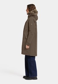 Braune Parkajacke mit fellgefütterter Kapuze, seitlichen Taschen und Druckknopfverschlüssen, kombiniert mit dunklen, weiten Jeans und schwarzen Plateau-Stiefeln.