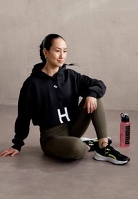 Felpa nera corta con stampa bianca a forma di 'H', leggings verde oliva, sneakers nere con dettagli gialli e una bottiglia d'acqua rosa accanto.