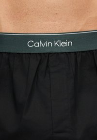 Μαύρα μποξεράκια Calvin Klein φορεμένα στη μέση, που δείχνουν υφή δέρματος και σκούρο ελαστικό ζωνάκι με το όνομα της μάρκας σε λευκά γράμματα.