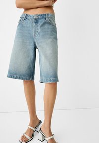 Person klädd i ljusblå denim Bermuda-shorts och vita högklackade sandaler, som står framför en helt vit bakgrund.