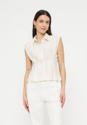 Ženska z dolgimi temnimi lasmi, oblečena v brezrokavno belo bluzo z naborki in bele hlače, stoji pred enostavnim svetlim ozadjem.