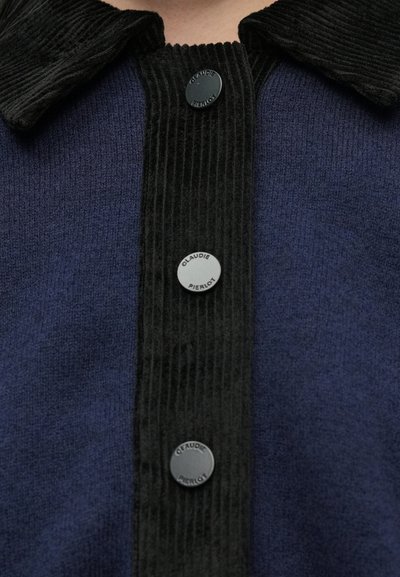 Μπλε σκούρα μπλούζα με γιακά και πατιλέτα από μαύρο κοτλέ ύφασμα. Διαθέτει στρογγυλά μαύρα κουμπιά με ανάγλυφη την επιγραφή "CLAUDIE PIERLOT". Μαλακή υφή.