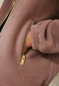 Zacht mauve fleece jack met een zijzak met een gouden rits en geribde manchetten, die een soepele textuur en een eigentijds ontwerp benadrukt.