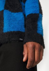 Sininen ja musta ruudullinen pörröneule, jossa on pehmeä pinta, varustettu ommellulla tarralla helmaosassa, päällä mustan kankaan.